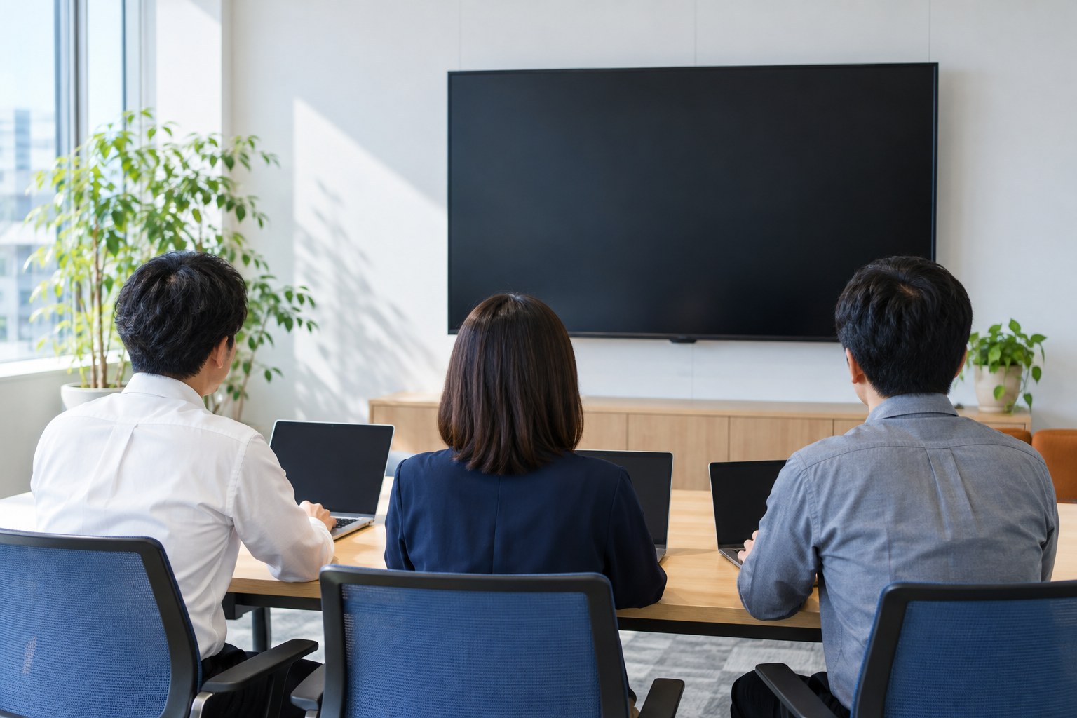 午前の会議室で、暗い大型モニターの前で打ち合わせる3人の後ろ姿